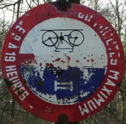 Photo d'un panneau interdisant aux vélos et motos de rouler sur un sentier pour piétons (Bois de la Houssière, ancienne voie du tram). La couleur s'est écaillée et l'on voit apparaître le panneau de stationnement alternatif sur lequel le «nouveau» panneau a été peint. Comble de malchance, la fixation du haut est rompue, et le panneau est maintenant retourné.